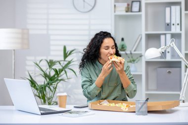 Modern bir ofis ortamında iş arasında masasında pizza yiyen bir kadın. Sahne, rahat bir anı sıradan bir yemekle yakalıyor..
