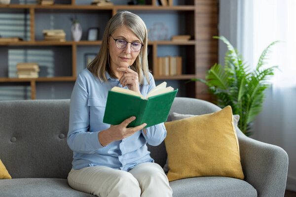 Elderly woman enjoying her reading time while sitting on a comfortable sofa at home. Relaxed and peaceful atmosphere with a book in hand.