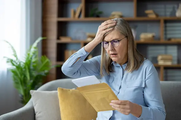 stock image Concerned elderly woman reading mail at home. The senior lady looks worried as she sits on a sofa holding an envelope.