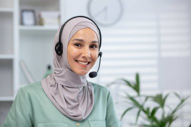 Başörtüsü ve kulaklık takan genç Müslüman kadının yakın plan portresi ofiste duruyor ve kameraya bakıp gülümsüyor..