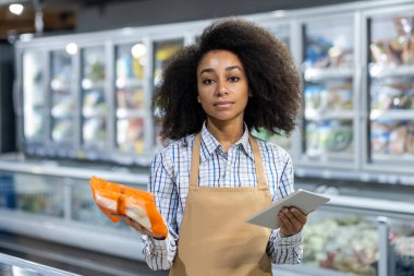 Süpermarkette çalışan genç bir Afro-Amerikalı kadının portresi buzdolabının yanındaki donmuş gıda bölümünde elinde bir tablet ve elinde bir paket yiyecek tutuyordu..