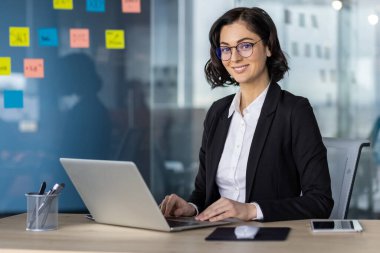 Modern ofis ortamında dizüstü bilgisayarda çalışan profesyonel bir kadın. Kendine güvenen ve odaklanmış, yapışkan notlar ve ofis malzemeleriyle çevrili..