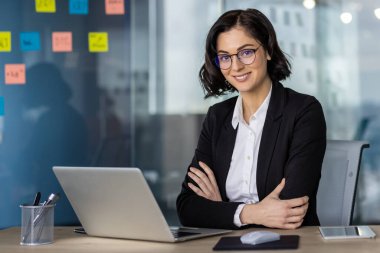 Ofis masasında oturan kendine güvenen bir iş kadını dizüstü bilgisayarı ve arka plandaki duvarda renkli yapışkan notlarla..