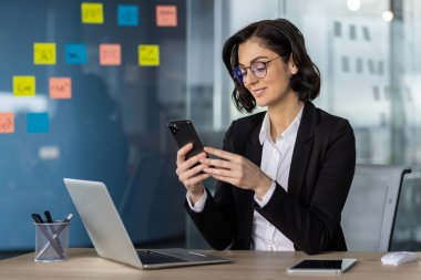 Modern bir ofis masasında bilgisayarlı akıllı telefon kullanan iş kadını, profesyonel çalışma ortamı ve çoklu görev kapasitesi gösteriyor.