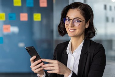 Modern bir ofiste akıllı telefon kullanan kendine güvenen bir iş kadını. Arka plandaki cam duvara yapıştırılmış notlar..