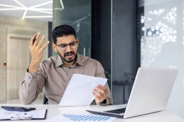 Gözlük takan genç bir adam ofis ortamında evrak tutarken stresli ve sinirli görünüyor. Elinde dizüstü bilgisayar ve belgelerle bir masada oturuyor. Endişe ve karmaşa hissi.