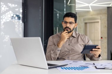 Odaklanmış bir finansör veya muhasebeci çağdaş bir ofiste dizüstü bilgisayardaki verileri analiz ederken bir hesap makinesi ve tablet kullanır. Düşüncelere dalmış, finans ve iş analizlerini ön plana çıkarıyor.