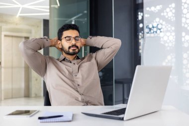 Kendine güvenen bir işadamı ofis masasında dinleniyor, işe ara veriyor. Bir dizüstü bilgisayar ve not defteri ile modern ofis alanı üretken ama dengeli bir çalışma ortamı yansıtır.