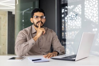 İşadamı, modern bir ofiste masasında çalışırken sessizliğini korumak için sessiz bir jest yapıyor..