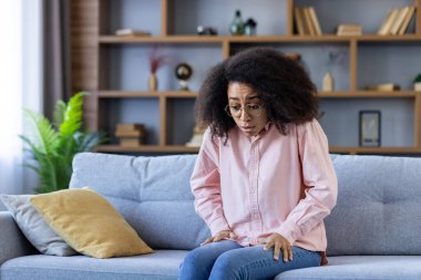 Kanepedeki kadın endişeli ve sıkıntılı görünüyor, muhtemelen panik atak geçiriyor. Kitaplık arka planlı modern oturma odası. Yüzünde anlamlı bir ifade olan pembe bir gömlek giyiyor..