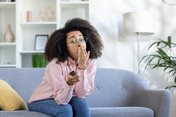 Woman yawning while watching TV at home, holding remote control on sofa. Boredom depicted in cozy home setting with modern interior design. Casual attire and relaxed environment