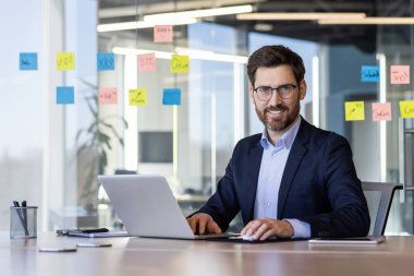 Modern ofis ortamında dizüstü bilgisayarda çalışan kendine güvenen bir iş adamı. Cam penceredeki yapışkan notlarla ofis ayarları. Gözlüklü, takım elbiseli, kameraya gülümseyen bir profesyonel..