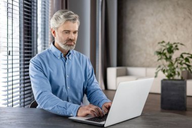 Gri saçlı, sakallı, modern ofis ortamında dizüstü bilgisayarla çalışan olgun bir adam. Ofis, pencereleri ve yeşil bitkileri olan minimalist bir tasarıma sahip, rahatlatıcı bir atmosfer yaratıyor.