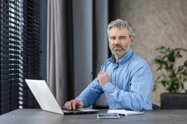Ev ofisinden dizüstü bilgisayar kullanarak çalışan kendinden emin olgun bir adam. Pencere perdelerinden doğal ışık saçan profesyonel bir ortam. Modern iş ortamı odak ve verimliliği yansıtıyor.
