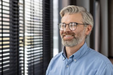 Çağdaş ofis ortamında panjurlarla pencereden dışarı bakarken gülümseyen gözlüklü olgun bir adam. Doğal ışık aydınlatma, neşeli ifade, profesyonel ama rahat tavır yakalama.