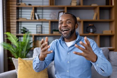 Kanepede oturup video görüşmesinde gülümseyen ve el kol hareketi yapan mutlu bir adam. Duygularını ifade eden ellerle canlı sohbete dalmak. Rafları ve bitkileri olan modern ev arkaplanı.