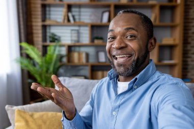 Evdeki kanepede otururken görüntülü konuşma yapan neşeli bir adam. Arkaplanda kitap rafı ve yanında yeşil bitki olan sıcak ve davetkar bir ortam. Dostça ve cana yakın bir hava yakalıyorum..