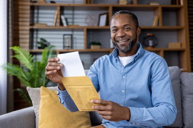 Adam rahat oturma odasında kanepede otururken mutlu bir şekilde zarfı açıyor. Kapalı alanda çekilen mutlu an, neşe, heyecan ve beklenti ifade eder..