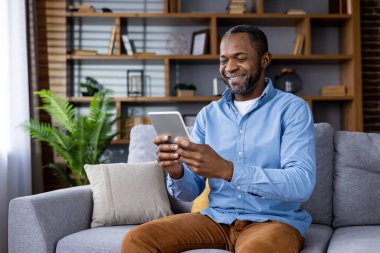 Gülümseyen adam rahat oturma odasında dijital tabletle etkileşime geçiyor. Rahat yaşam tarzı, teknoloji kullanımı ve ev konforu. Ahşap rafları ve dekoratif bitkileri olan modern oturma odası.