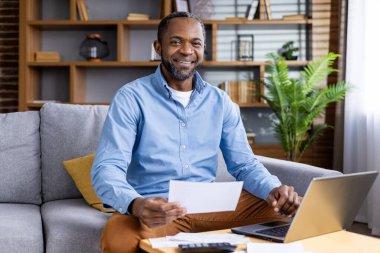 Adam elinde belgelerle çalışırken gülümsüyor. Laptop ve kırtasiyeyle ev ofis ortamında rahat ve üretken bir atmosfer..