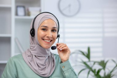 Gülümseyen kadın müşteri hizmetleri temsilcisi kameraya bakarken kulaklık takıyor. Profesyonel çevre, modern ofis düzeni, kendine güvenen ve ulaşılabilir. İletişim kavramı, destek