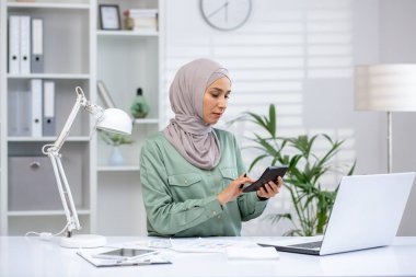 Kadın muhasebeci, modern ofis masasında dizüstü bilgisayar ve belgelerle çalışırken verileri hesap makinesiyle hesaplar. Ekonomide doğruluğa odaklanmak.