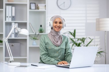 Başörtüsü takan gülümseyen bir kadın ev ofisinde kulaklıkla dizüstü bilgisayarla çalışıyor. Beyaz mobilyalar, kitaplık ve bitkilerin yer aldığı parlak ve modern çalışma alanı. Uzak çalışma ve üretkenlik kavramı
