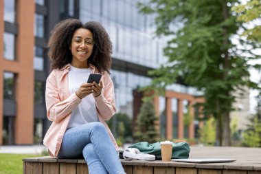 Üniversite kampüsünün yakınında oturan genç bir kız öğrenci akıllı telefon kullanıyor, elinde kahve fincanı ve sırt çantasıyla tahta güvertede güneşli bir günün tadını çıkarıyor, günlük giysiler giyiyor ve gülümsüyor.