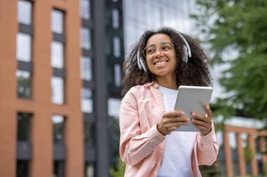 Üniversite kampüsünün dışında tablet kullanan kulaklıklı mutlu genç kız öğrenci. Öğrenci modern eğitim ortamında öğrenim ve müzikten hoşlanıyor.