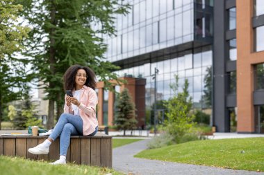Üniversite kampüsünün yakınında oturan genç bir kız öğrenci elinde akıllı telefon tutuyor. Gündelik kıyafetler giyen gülümseyen kadın, arka planda modern binalar olan kampüste açık havanın tadını çıkarıyor. Rahatlatıcı ve