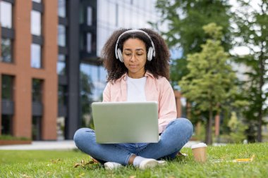 Üniversite kampüsünün yakınında çimlerde oturan genç bir kız öğrenci kulaklıkla dizüstü bilgisayarla çalışıyor. Doğal ortamda çalışmak ve teknolojinin tadını çıkarmak için güzel bir gün..
