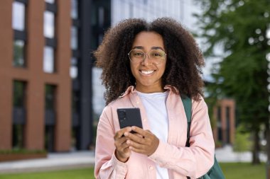 Üniversite kampüsünün yanında elinde akıllı telefon ve gülümsemeyle duran mutlu genç kız öğrenci. Parlak güneşli bir gün, arka planda modern binalar, günlük kıyafetler, öğrenim, eğitim