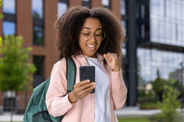 Gözlüklü ve sırt çantalı genç bir kız öğrenci üniversite kampüsünün yakınındaki akıllı telefonlara bakarken iyi haberleri kutluyor. Arka planda modern bina olan açık hava sahnesi.
