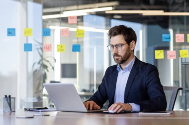 Modern ofisteki profesyonel iş adamı, cam duvarda yapışkan notlarla dizüstü bilgisayarda çalışıyor. Çağdaş çalışma alanındaki verileri analiz eden takım elbiseli ve masasında gözlüklü adam..