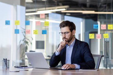 Modern ofiste dizüstü bilgisayarda çalışan bir iş adamı ve cam duvarda yapışkan notlar var. Takım elbise analizi, beyin fırtınası ve iş başarısı stratejisi üzerinde odaklanmış bir profesyonelsin..