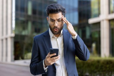 Şok olmuş mavi takım elbiseli işadamı, modern ofis binasının dışında şaşırmış bir ifadeyle akıllı telefona bakıyor. Şaşırmış, stresli ve endişeli hissederken beklenmedik haberler veya hatalar okuyor..