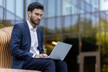 Profesyonel kıyafetlerle dışarıda dizüstü bilgisayarda çalışan bir iş adamı. Arka plandaki modern ofis binası. Profesyonel, odaklanmış ve işiyle meşgul. Verimliliği ve modern çalışma ortamını temsil eder