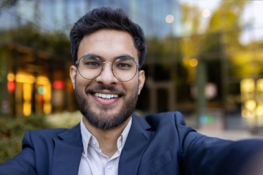 İş adamı gülümsüyor ve gözlük takıyor ve dış görünüşü bulanık modern binalar ve yeşillikler sergiliyor.