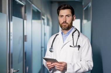 Steteskoplu beyaz önlüklü kendine güvenen doktor elinde tabletle klinik koridorunda dikiliyor. Tıbbi uzman hasta muayenesi için hazır. Modern tıbbi tesiste sağlık işçisi.