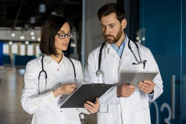 Doktorlar hastane koridorunda hastaların sağlık raporlarını tartışıyorlar. Elinde tablet olan erkek doktor, elinde not panosu olan kadın doktor. Tıp uzmanları işbirlikçi sağlık hizmetleri değerlendirmesi yaptılar.