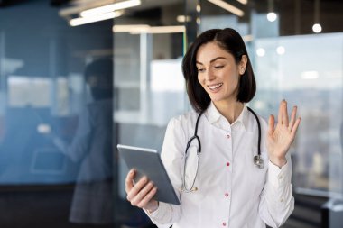 Genç bayan doktor, modern ofiste video araması için tablet kullanıyor. Steteskoplu beyaz önlüklü profesyonel sanal danışmanlık yapıyor, hastalarla çevrimiçi bağlantı kuruyor, ekranda el sallıyor.