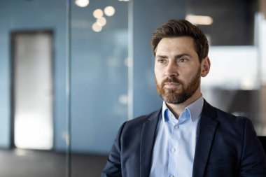 Modern ofis ortamında sakallı ve takım elbiseli profesyonel iş adamı özgüvenli ve düşünceli görünüyor. Profesyonel davranışı vurgularken yumuşak odak arkaplanı derinliği ekleme