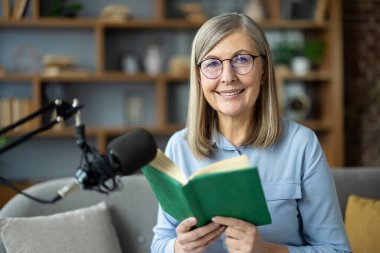 Evde podcast yapan yaşlı bir kadın, açık bir kitap tutuyor ve kameraya gülümsüyor. Gözlük takıyor ve rahat bir oturma odasında oturmuş podcast 'ini kaydediyor..
