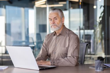 Modern ofis ortamında dizüstü bilgisayarı üzerinde çalışan tecrübeli bir iş adamının portresi. Düşünceli ve odaklanmış, işyerinde üretkenlik ve adanmışlığı somutlaştırıyor..