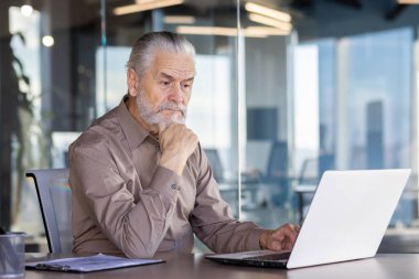 Üst düzey işadamı, modern ve iyi aydınlatılmış bir ofis ortamında dizüstü bilgisayar üzerinde çalışıyor. Çalışırken odaklanmış ve derin düşüncelere dalmış görünüyor. Bu görüntü profesyonelliği ve tecrübeyi aktarır..