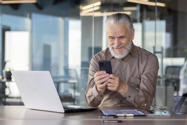 Modern ofis ortamında akıllı telefon kullanırken kıdemli işadamı gülümsüyor. Elinde dizüstü bilgisayar, gözlük ve evrak işleriyle bir masada oturuyor. Bu da profesyonel görevlerde aktif rol aldığını gösteriyor..
