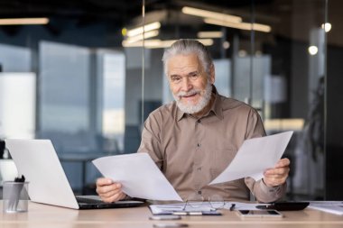 Modern bir ofiste çalışan kıdemli işadamı elinde gülümseyerek belgeleri tutuyor. Görüntü profesyonelliği, deneyimi ve bir dizüstü bilgisayar ve çalışma alanına sahip kurumsal bir ortama olan güveni resmediyor.