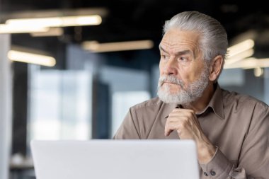 Çağdaş bir ofis ortamında dizüstü bilgisayarla çalışan düşünceli bir ifadeye sahip yaşlı bir işadamı. Görüntü profesyonelliği, deneyimi ve derin tefekkürü aktarır.,