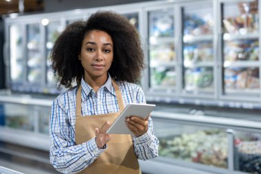 Market koridorunda duran genç bayan çalışan, envanter kontrolü için dijital tablet kullanıyor. Önlüklü sıradan bir kıyafet giyiyor.