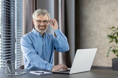 Kendine güvenen orta yaşlı bir işadamı gözlüklü ve mavi gömlekli modern ofis ortamında dizüstü bilgisayarıyla çalışırken gülümsüyor. Görüntü profesyonelliği, pozitifliği ve üretkenliği yansıtıyor.